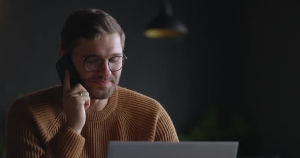 Κοντινό πλάνο του ευτυχισμένου άντρα να μιλάει στο κινητό στο γραφείο. Πορτρέτο του χαμογελαστού ανθρώπου που εργάζεται σε φορητό υπολογιστή στην καφετέρια και μιλώντας στο smartphone — Αρχείο Βίντεο