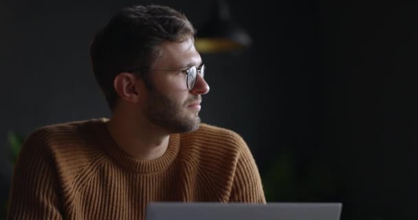 Eine Nahaufnahme zeigt einen nachdenklichen, ernsthaften Mann in Brille, der mit einem Laptop im dunklen Büro über Lösungen nachdenkt. Zeitlupe — Stockvideo