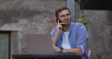 Mavi tişörtlü yakışıklı bir adamın orta boy fotoğrafı dizüstü bilgisayarda çalışırken ve kafede otururken cep telefonuyla cevap verirken. Yavaş çekim