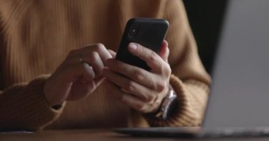 Dizüstü bilgisayar başında çalışırken cep telefonu kullanarak saatli erkek ellerini yakın plan çek. Erkek parmaklar akıllı telefondan yazıp dinliyorlar. Mesaj ve kaydırma. Yavaş çekim.