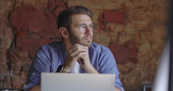 Pensativo hombre serio que lleva en camisa azul en gafas sentarse con el ordenador portátil pensando en la solución en la cafetería y mirando a la ventana. Movimiento lento — Vídeos de Stock