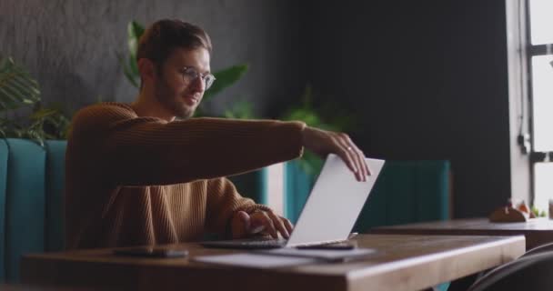 Hezký muž v brýlích v hnědém svetru dokončí práci v laptopu a zavře ho. Na volné noze sedí v kavárně a pracuje na notebooku. Zpomalený pohyb — Stock video