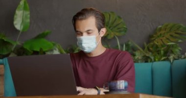 Koruyucu maskeli bir adam COVID sırasında rahat bir kafede kahve içiyor. Genç adam dizüstü bilgisayarında kayıt yapıyor.