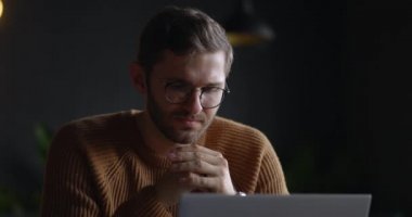 Gözlüklü düşünceli, ciddi bir adam laptopa bakıyor ve solüsyonu karanlık ofiste düşünüyor. Yavaş çekim