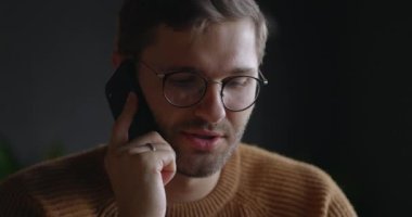 Gözlüklü adamın karanlık arka planda konuşan cep telefonunun yakın görüntüsü. İş adamının iş yerinden ağır çekimde telefon görüşmesi yapması. Telefon eden iş adamının portresi.