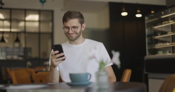 Hombre guapo en gafas con una camiseta blanca utiliza un teléfono inteligente y sonríe en la cafetería. Hombre alegre navegando por Internet por teléfono móvil mientras pasa tiempo libre en un acogedor café. — Vídeos de Stock