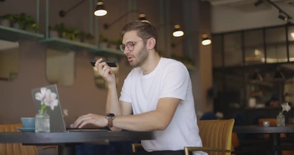 Seitenansicht eines gutaussehenden, ernsten Mannes mit Brille und weißem Hemd, der am Tisch im Café sitzt und per Laptop Sprachnachrichten per Smartphone verschickt. Zeitlupe — Stockvideo