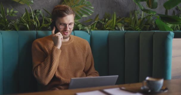 갈색 스웨터를 입고 안경을 쓴 남자가 휴대폰으로 이야기하고 카페에서 노트북을 사용하고 있습니다. 원격 작업의 개념. 느린 동작 — 비디오