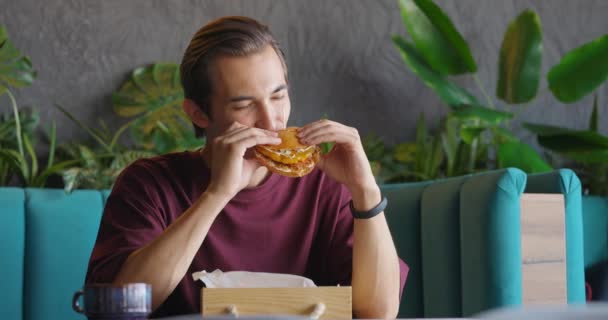 Giovane uomo che indossa una maglietta godendo di hamburger in caffetteria. Ragazzo che prende una tazza di caffè durante il pranzo — Video Stock