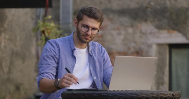 Estudante inteligente vestindo óculos e camisa azul estudando com laptop e beber café gelado no café ao ar livre. Educação remota em e-learning. Movimento lento. — Vídeo de Stock