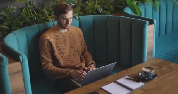 Πλευρική άποψη του ανθρώπου που κάθεται στον καναπέ χρησιμοποιώντας το φορητό υπολογιστή για την απομακρυσμένη εργασία. Όμορφος ελεύθερος επαγγελματίας επιχειρηματίας με γυαλιά που δουλεύει στο καφέ. Σπουδαστές online — Αρχείο Βίντεο