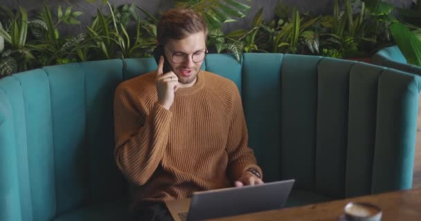 갈색 스웨터를 입고 안경을 쓴 남자가 휴대폰으로 이야기하고 카페에서 노트북을 사용하고 있습니다. 원격 작업의 개념. 느린 동작 — 비디오