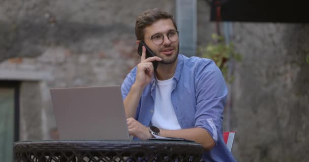 Mavi tişörtlü bir adam dizüstü bilgisayarla çalışıyor ve sokak kafesinde otururken cep telefonuyla konuşuyor. Yavaş çekim — Stok video