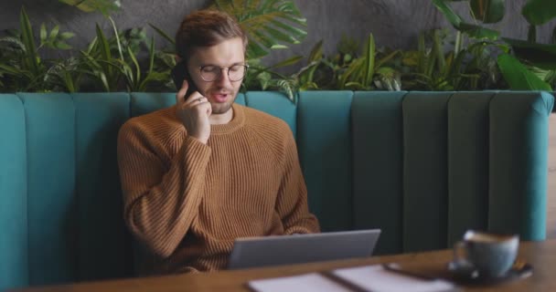 Muž v hnědém svetru a brýlích sedí na pohovce, mluví na mobilu a pracuje na notebooku v kavárně. Zpomalený pohyb — Stock video