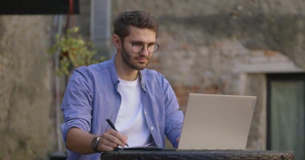 Estudiante inteligente con anteojos y camisa azul estudiando con portátil y escribiendo en cuaderno en la cafetería al aire libre. Educación electrónica remota. Movimiento lento. — Vídeos de Stock