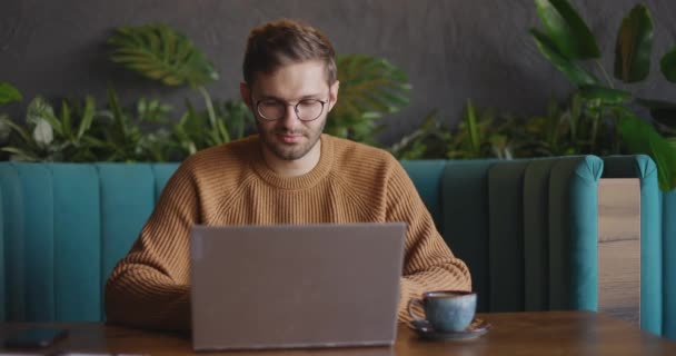 Ένας ενήλικας κάθεται στο καφέ την ημέρα και χρησιμοποιεί το λάπτοπ για την απομακρυσμένη δουλειά. Όμορφος ελεύθερος επαγγελματίας επιχειρηματίας με γυαλιά που δουλεύει στο καφέ. Σπουδαστές online. — Αρχείο Βίντεο