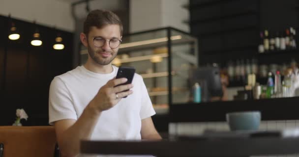 Attraente uomo serio in occhiali da vista indossa in camicia bianca seduto al tavolo al caffè e utilizzando smartphone. Messaggi di testo che lavorano guardando video di routine quotidiana divertente comunicare — Video Stock