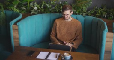 Kahverengi kazaklı ve gözlüklü yetişkin adam gündüz vakti kanepede oturuyor ve uzaktan kumanda için dizüstü bilgisayarı kullanıyor. Yavaş çekim