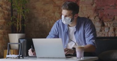 Maskeli genç bir iş adamı dizüstü bilgisayar kullanıyor ve kahve dükkanında oturuyor. Koruyucu maske takan yakışıklı bir öğrenci. Uzaklık eğitimi