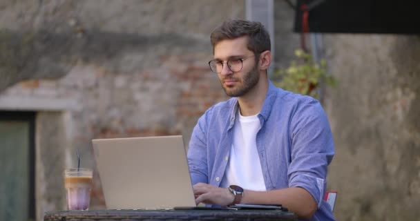Foto seorang pemuda berbaju biru duduk di meja di kafe terbuka atau ruang kerja bersama dan menggunakan komputer laptop. Gerakan lambat — Stok Video