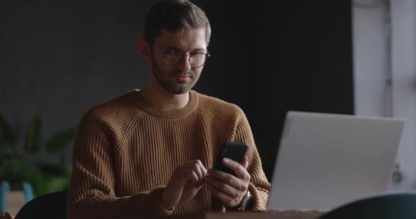Seorang pria dengan sweater coklat dan kacamata menggunakan ponsel dan laptop saat duduk di kedai kopi. Bisnis, teknologi, komunikasi, pendidikan. Gerakan lambat — Stok Video