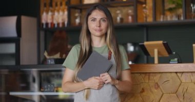 Kafede ya da restoranda elinde menü defteriyle kameraya bakan önlüklü güzel garson kız. Yavaş çekim.