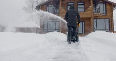 Bir adam kar yağışı sırasında ahşap evin yanında kar temizleme makinesiyle kar temizliyor. Arkadan bak. Yavaş çekim