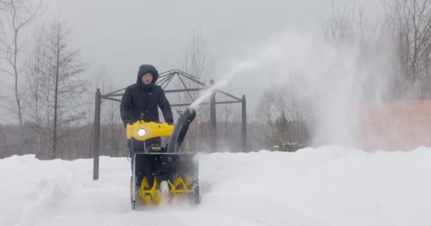 Een man reinigt de sneeuw met een sneeuwblazer in de tuin bij sneeuwval in slow motion. Vooraanzicht. Langzame beweging — Stockvideo