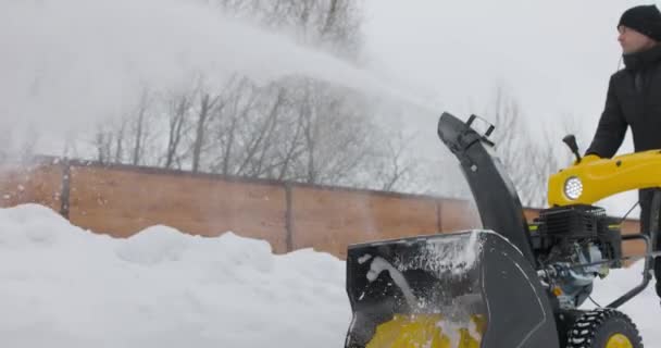 Een man die met een sneeuwblazer werkt. Sneeuwwitje winter. De mens verwijdert sneeuwvlokken met sneeuwploeg. Sneeuwblazer in actie. Koud weer. Langzame beweging — Stockvideo