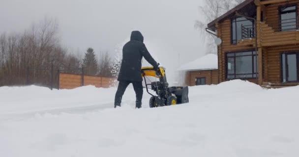 Человек чистит снег снегоочистителем задний план деревянного дома в зимний период. Вид сзади. Медленное движение — стоковое видео