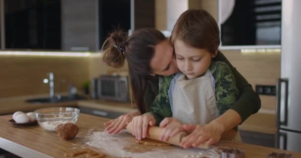 Mladá rodinná matka a syn dělají doma upečené sušenky u kuchyňského stolu a pracují s těstem a usmívají se. Matka líbá dítě. Zpomalený pohyb — Stock video