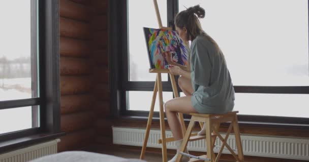 Hobby en casa durante el auto-aislamiento, artista mujer está pintando cuadro sobre lienzo cerca de la ventana en casa de madera. Movimiento lento — Vídeo de stock