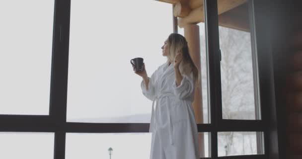 Joven hermosa mujer en albornoz blanco beber café junto a la ventana en casa por la mañana — Vídeos de Stock