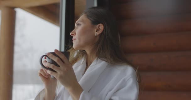 Evinin penceresinin yanında bardağı olan güzel ve sağlıklı bir kadın. Sabahları sıcak kahve içen bornozlu mutlu kadın.. — Stok video