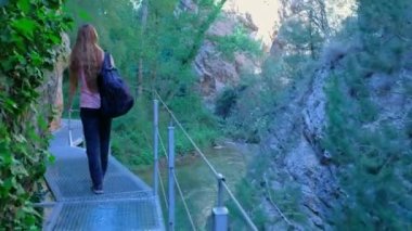 Woman is crossing a river on a hanging bridge with a backpack. Girl is hiking in nature. Green forest landscape. Horizontal video.