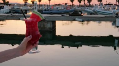 Gün batımında deniz kenarında taze karpuzla süslenmiş tropik bir kokteyli tutan kadın. Arka planda tekneler olan liman. Yaz tatili havası. Tam HD video. 