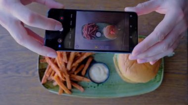 Adam yemek fotoğrafı çekiyor. Patates kızartmalı lezzetli bir hamburger. Tahta bir masada taze pişmiş lezzetli burger. Sebzeli burger. 