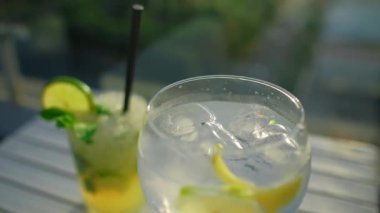 Taze limon ve nane yapraklarıyla süslenmiş tropik kokteyl. Mojito ve cin tonik. Arka planda güzel bir günbatımı. Yaz tatili havası. Tam HD yavaş çekim videosu. Arkaplanda gün batımı.
