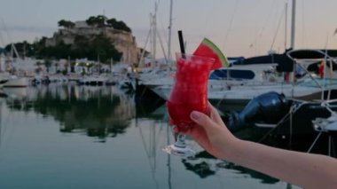 A hand with a tropical cocktail decorated with fresh watermelon at the seaside. Harbour with boats on the background. Vacation summer vibe. Full HD video. 