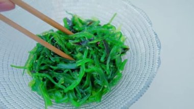 Close up of an algae with chopsticks. Soaked green wakame seaweed in a glass bowl. Japanese food. Healthy seafood. Slow motion video. Isolated on white. Top view. 