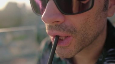 Adam kokteyl içiyor. Güneş gözlüklü ve pipetli bir suratı kapat. Yavaş çekim videosu. Yavaş çekim videosu. Arkaplanda gün batımı. 