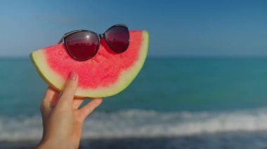 A hand holding a watermelon with sunglasses. Blue turquoise Mediterranean sea with waves on the background. Slow motion video. 