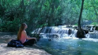 Kadın şelaleli bir doğada oturuyor ve meditasyon yapıyor. Şelalesi olan güzel bir manzara. Yeşillik yapraklı yeşil orman. Güneşli güzel hava. Yatay video. 