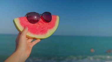 A hand holding a watermelon with sunglasses. Blue turquoise Mediterranean sea with waves on the background. Slow motion video. 