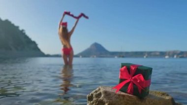 Uzun saçlı, kırmızı bikinili ve Noel Baba şapkalı bir kadın. Kız mutlu ve havaya şapka fırlatıyor. Arka planda Akdeniz. Mavi gökyüzü ve dağlar.