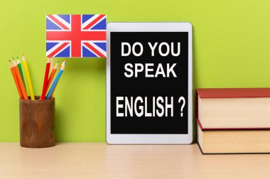 A digital tablet with the text do you speak english? written in it, a pot with pencils and the flag of the United Kingdom, on a wooden desk, with green background