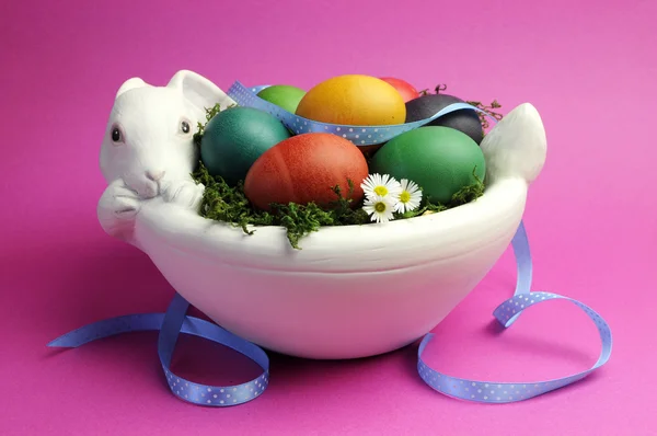 Rainbow color easter eggs in white bunny rabbit bowl with blue polka dot ribbon on a pink background. — Stock Photo, Image