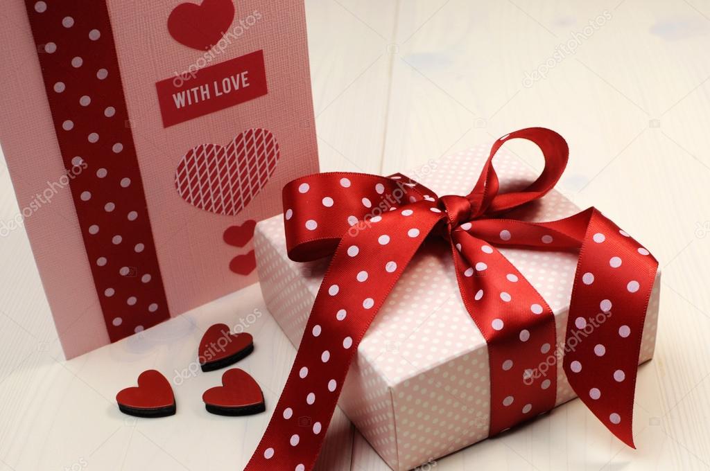 Handmade gift card with pink gift and red polka dot ribbon and heart on white natural wood table.