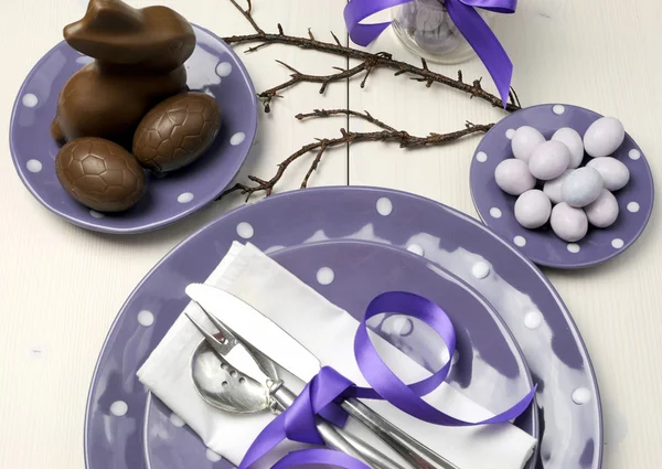 Purple theme Easter dinner, breakfast or brunch table setting with chocolate bunny rabbit, and sugar candy coated birds nest eggs on polka dot plates on white shabby chic natural wood table. — Stock Photo, Image