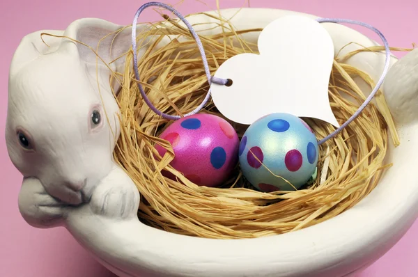 Pink and blue Easter eggs in white bunny bowl with heart gift tag on pink background. — Stock Photo, Image